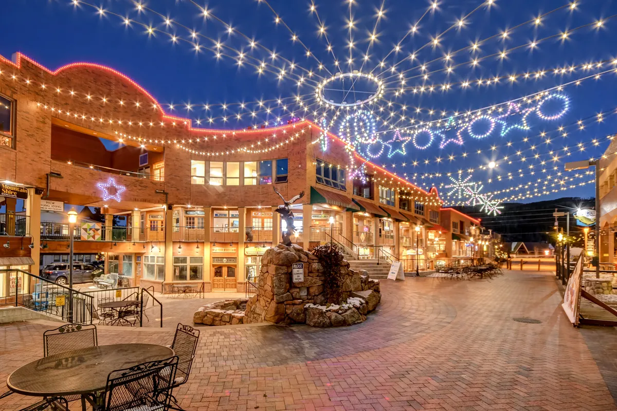 A vibrant exterior scene showcasing decorative storefronts adorned with string lights, a central fountain, and people enjoying a festive atmosphere. The nighttime lighting and cheerful decor evoke the fun of shopping, dining, and community gatherings in the town center.