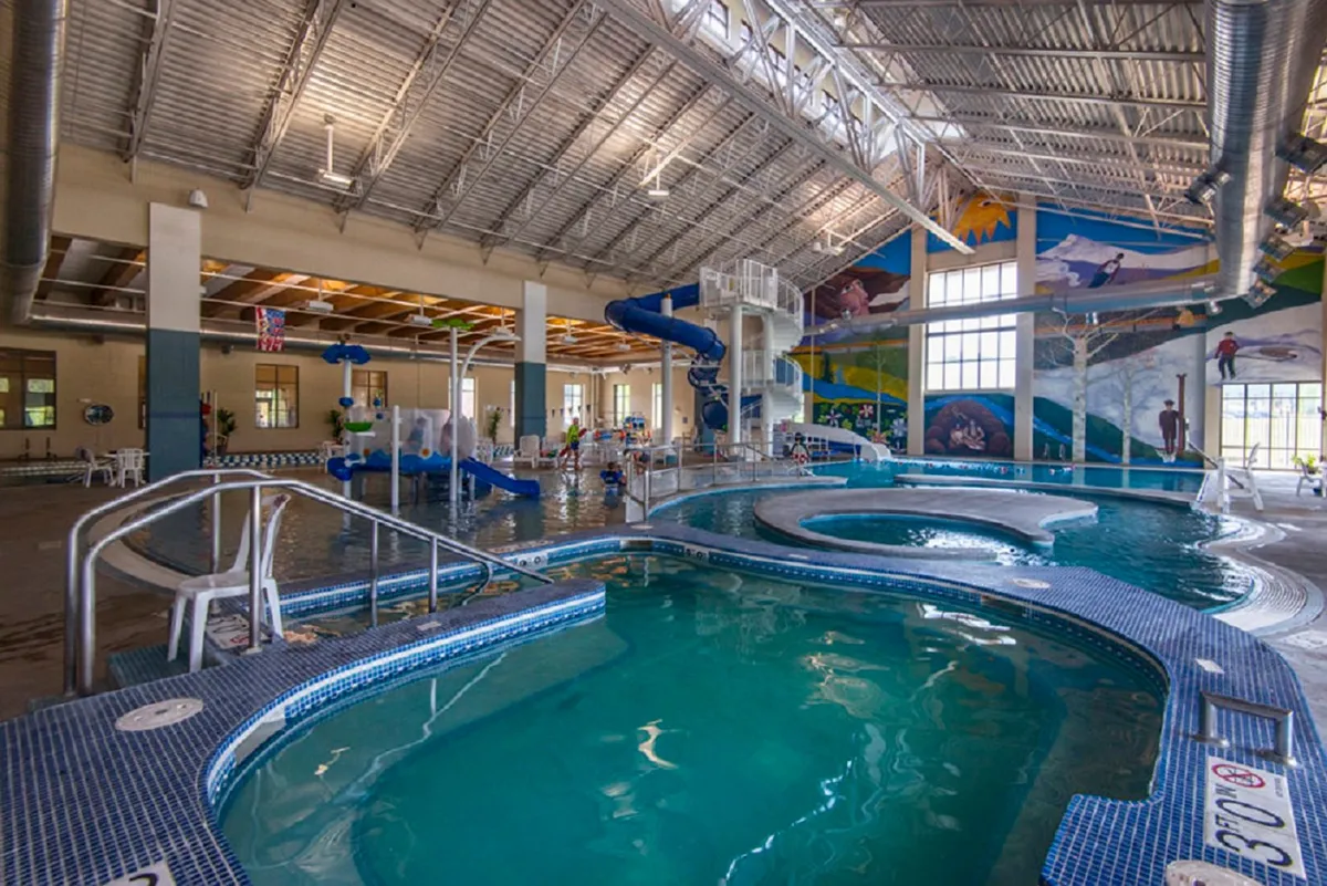 A lively indoor recreation center scene featuring a swimming pool, waterslides, a mural on the wall, and children playing. The atmosphere is joyful and bustling with family activity.