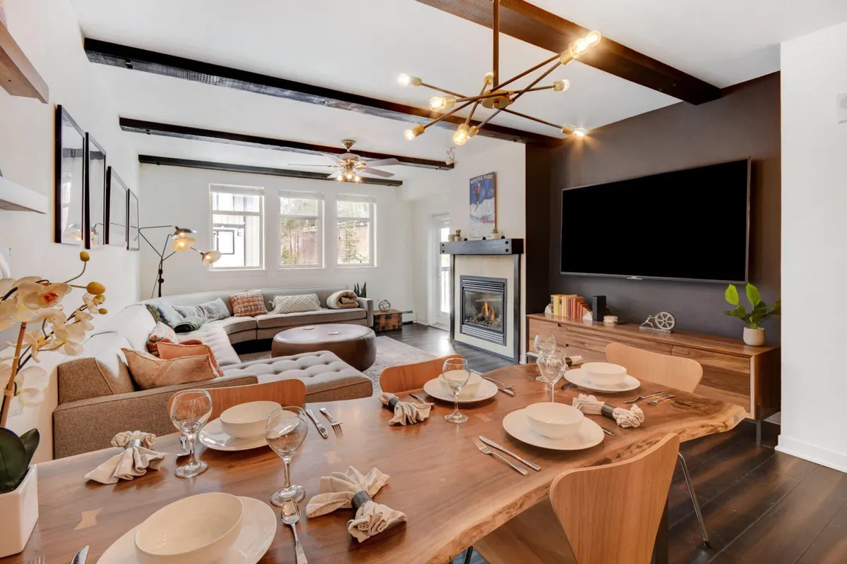 This image features a modern, cozy living room with a plush sofa, a stone gas fireplace, a large HDTV, and an adjacent dining table. Expansive windows provide ample natural light, suggesting sweeping views just beyond, and the inviting arrangement encourages relaxation and social gatherings.