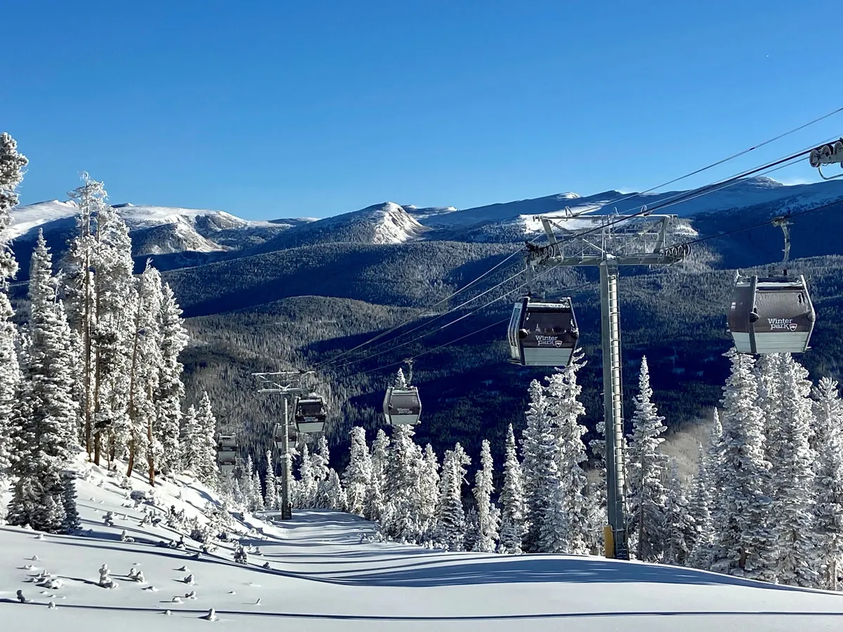 A striking exterior view featuring a ski lift ascending over snow-covered trees, with a sweeping mountain range in the background. The pristine white snow and clear blue sky evoke the thrill and beauty of alpine skiing. The ski lift is in motion, and the perspective provides a sense of adventure and direct access to the slopes.