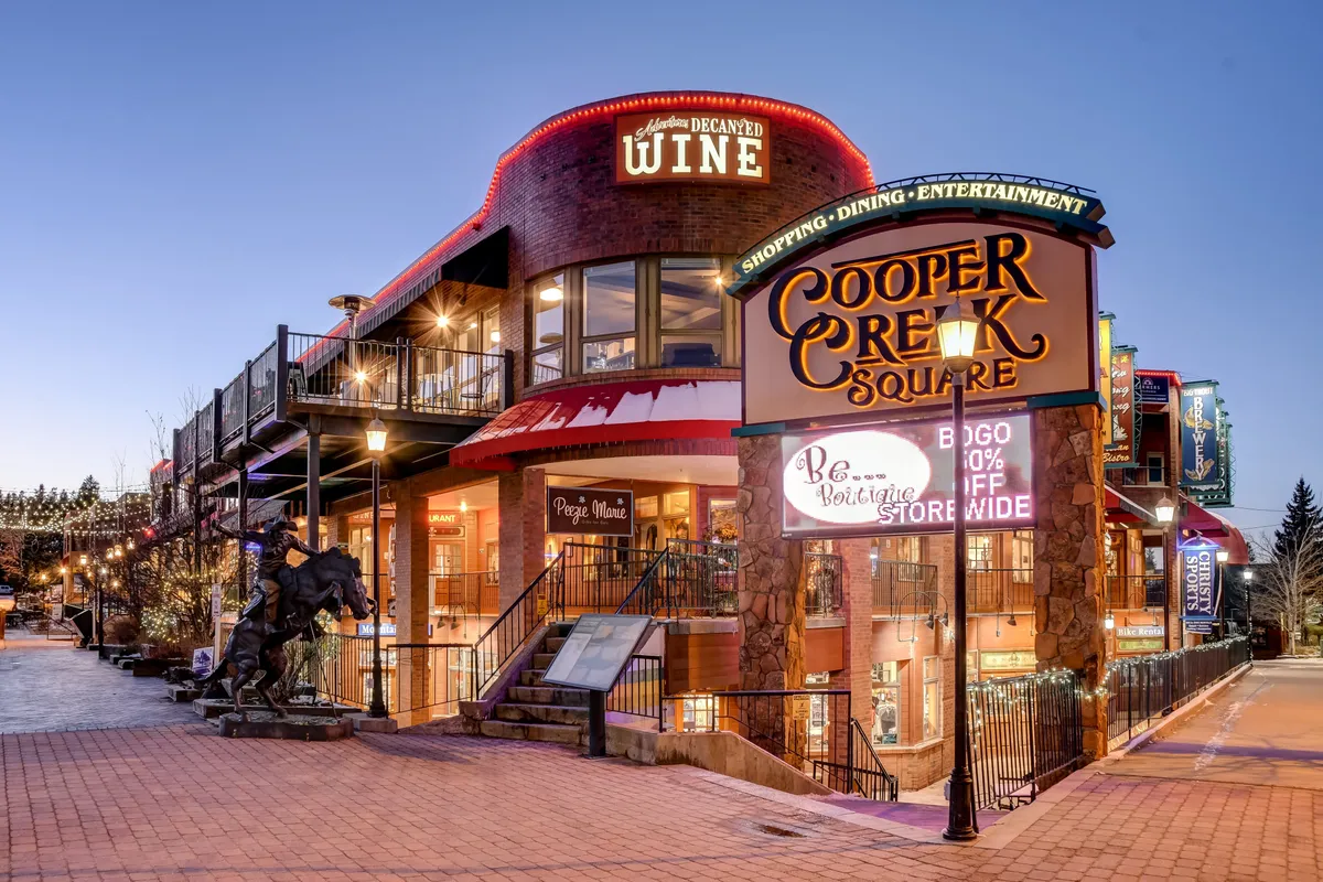 A vibrant exterior of Cooper Creek Square, featuring welcoming signage, a wine shop, outdoor seating, and festive string lights. The scene is lively and inviting, with hints of dining and shopping opportunities and a backdrop of the open, bustling town center.
