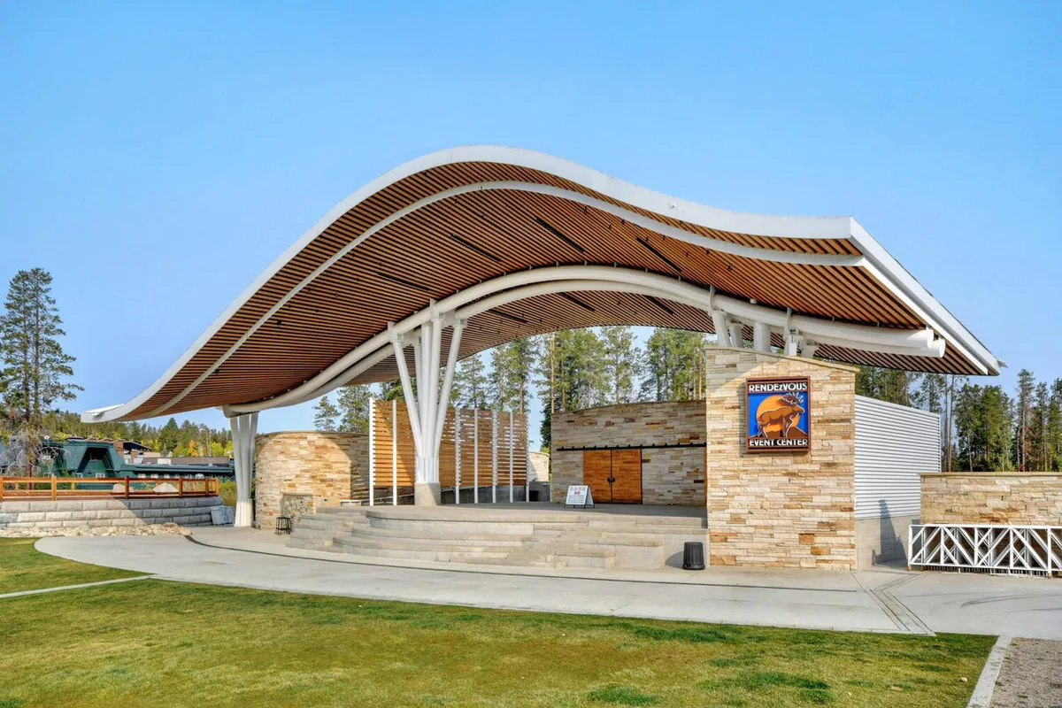 An outdoor amphitheater with a distinctive curved roof and stone facade, set amidst a welcoming plaza. The stage area is visible, with ambient lighting suggesting a setting for community events, concerts, or festivals. The environment is festive and inviting, hinting at year-round outdoor entertainment.