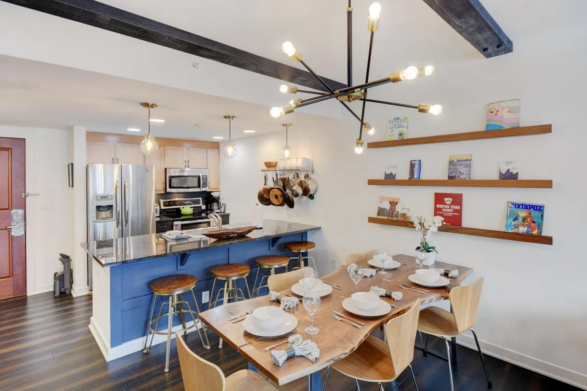 A bright, modern open-plan kitchen and dining area featuring a dining table set for a meal, a kitchen island with bar stools, shelves with books and decorations, and a striking modern light fixture. The space looks ready for family gatherings or festive dinners, with a stylish yet cozy atmosphere.