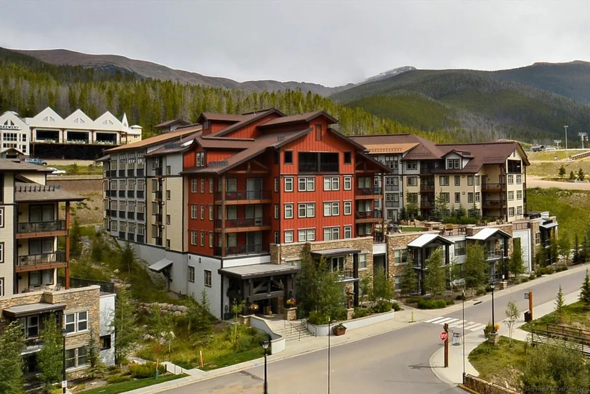 An exterior view of a central building with red accents surrounded by lush greenery and a dramatic mountain backdrop. The scene feels inviting and tranquil, suggesting easy access to outdoor trails and activities right from the doorstep.