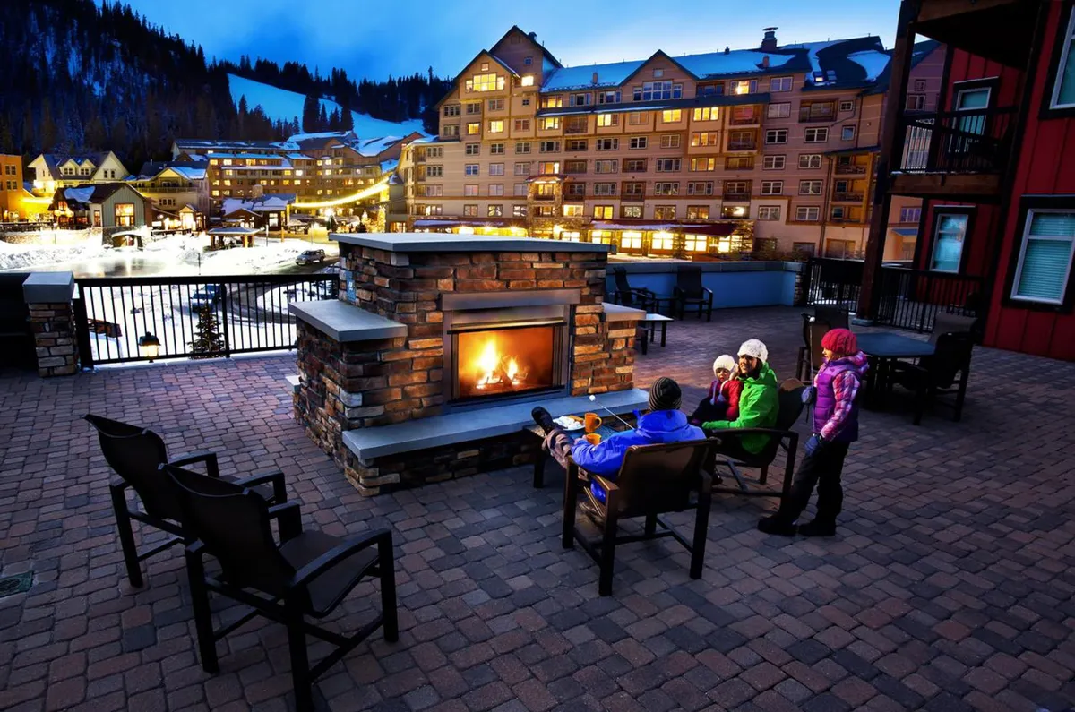 A welcoming exterior lounge area featuring a double-sided outdoor fireplace, ample seating, and a dramatic mountain backdrop. The area is illuminated for evening gatherings and conveys a sense of warmth and camaraderie.