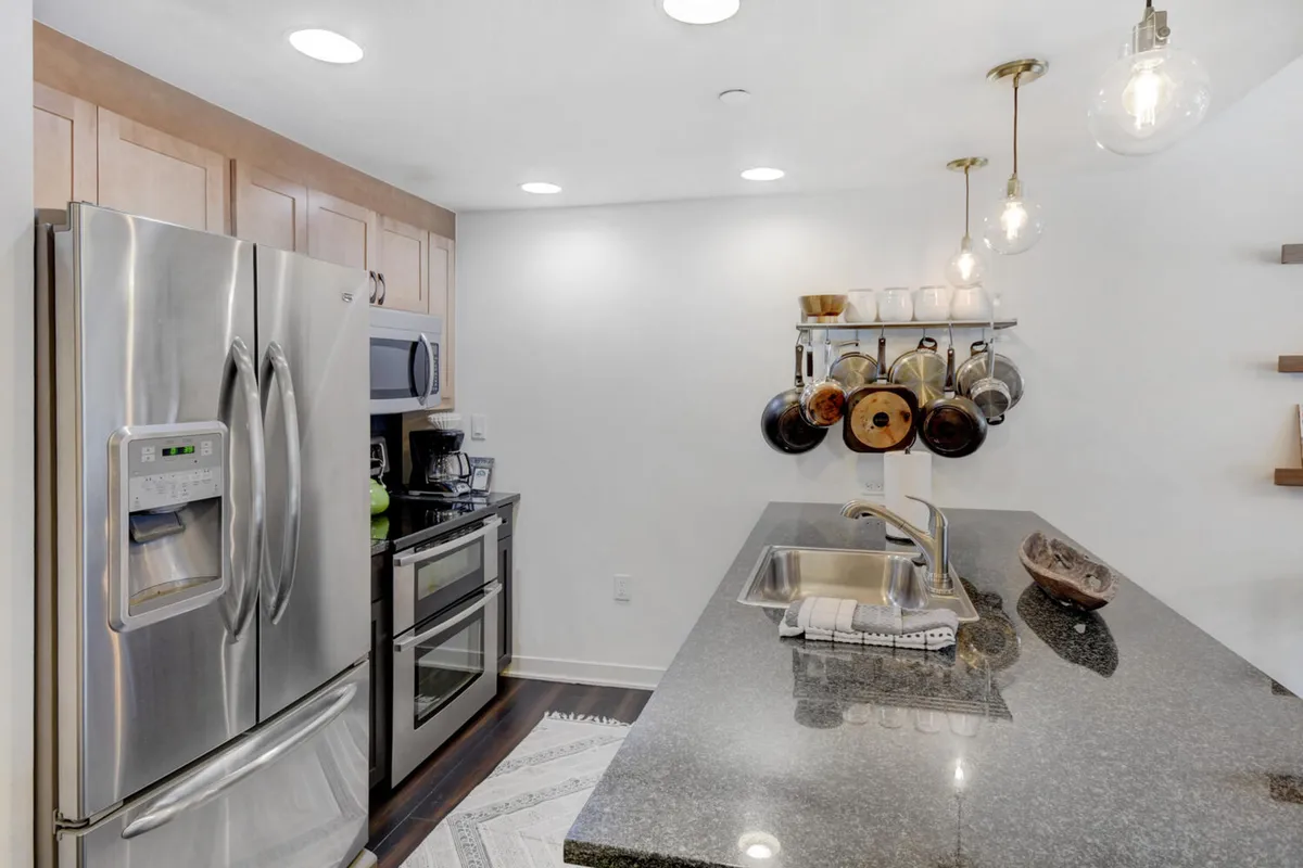 A kitchen scene focused on high-end stainless steel appliances, a granite countertop, and hanging pots and pans above a wooden shelf with dishes. The space is functional and inviting, clearly equipped for gourmet cooking and family meals.