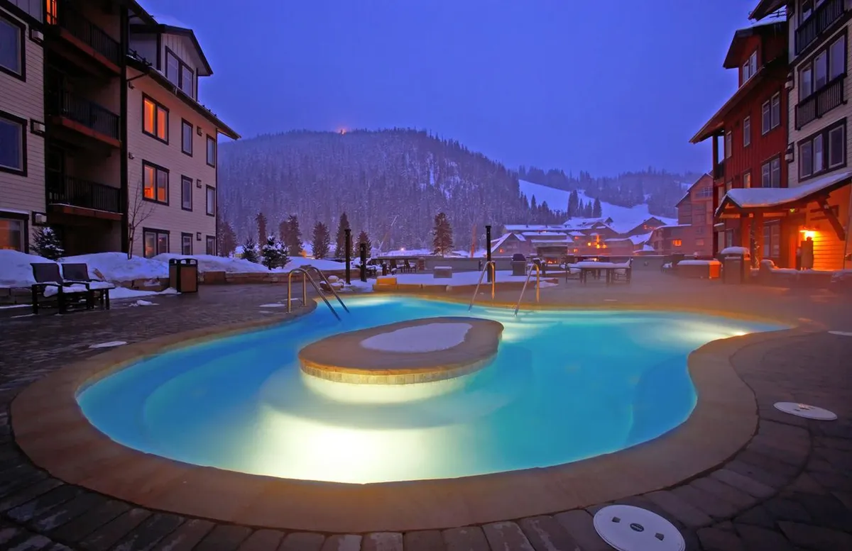 A serene outdoor hot tub area surrounded by snow, with an illuminated pool and views of the mountains in the background. The ambiance is tranquil, suggesting relaxation and socializing after a day of adventure.